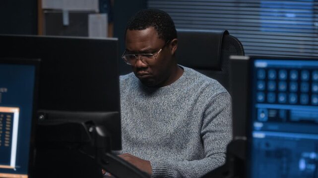 Experienced Black male IT security professional analyzing digital data on multiple screens in focused office environment while monitoring network and system security