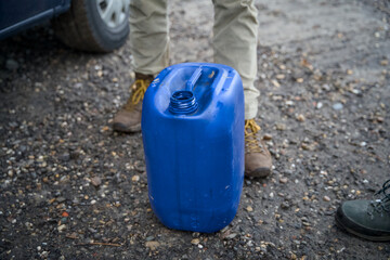 Open blue jerrycan with visible contents, placed on the ground in front of a pair of work boots. Practical storage container in a farm or workshop setting.