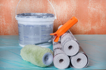 Wallpaper rolls, glue, paint, a roller, and a brush are ready for an apartment renovation. Room renovation tools are displayed against an old wall. Apartment renovation concept