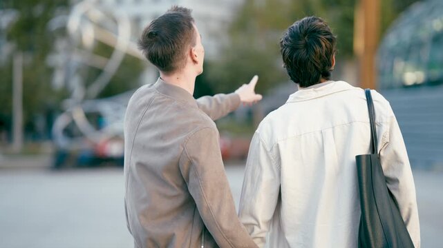 Back view of a young gay couple holding hands and walking together in a city. One man points at something while they talk and enjoy their date