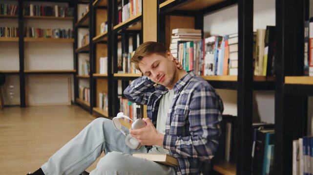 Young tired man sits on the floor of a library enjoying a book while listening to music with headphones in a quiet corner during the afternoon