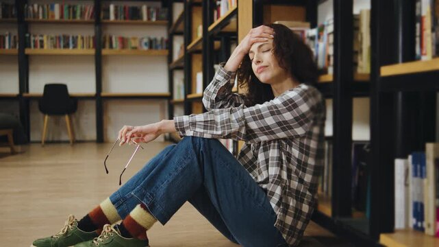 Tired woman sitting on the floor of a library looking stressed while holding glasses in a cozy corner during daytime