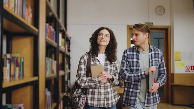 Two students walk in a library while discussing a project during the afternoon with books and study materials around them