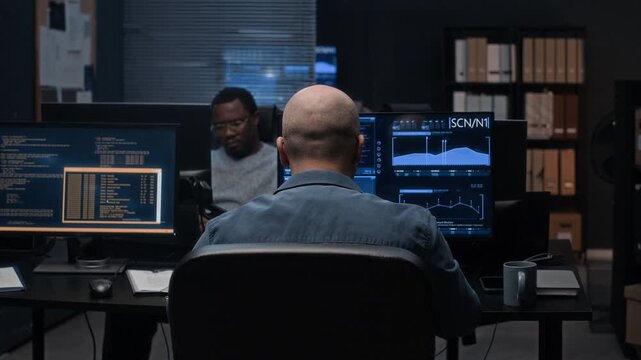 Back view shot of bald cybersecurity analyst working late in dark office reviewing multiple screens with Black male colleague sitting in background, analyzing data and monitoring security systems