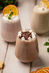 Creamy chocolate drinks topped with marshmallows and orange slices served in glasses on a rustic wooden table