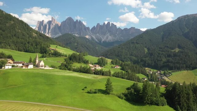 Aerial view of a quaint village nestled amid verdant hills and towering mountains, a church spire punctuating the skyline, Funes, Trentino-Alto Adige, Italy.
