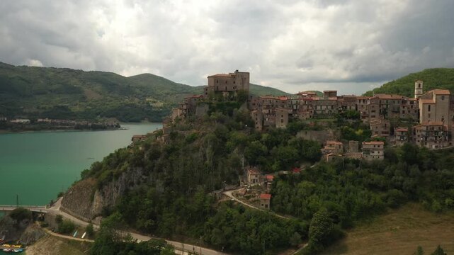 Aerial view of the ancient village of Turano clinging to a rocky spur overlooking the serene blue-green waters of Lake Turano, Turano, Lazio, Italy.