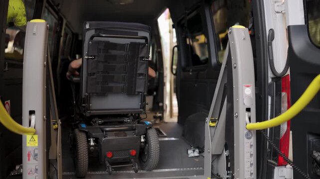 Man operating a hydraulic lift with a remote control, helping a person in an electric wheelchair enter an accessible van for transport
