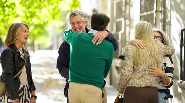 Mature son happily greeting and hugging his visiting senior parents and their friends on a sunny city sidewalk during a reunion