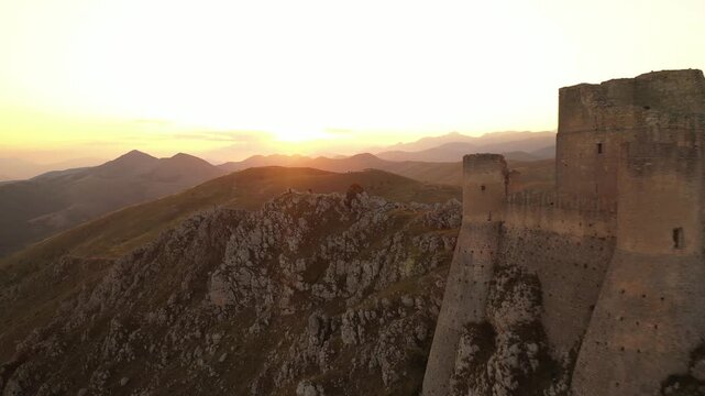 Aerial view of Rocca Calascio, a mountaintop fortress, bathed in the warm, golden light of the setting sun, creating a stunning contrast with the rugged landscape, Calascio, Abruzzo, Italy.