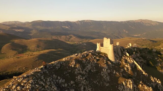 Aerial view of the Rocca Calascio fortress perched high on a hilltop, bathed in golden sunlight, contrasting with the rugged terrain, Calascio, Abruzzo, Italy.