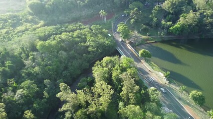 Aerial view of a road and bridge beside an urban lake, surrounded by green vegetation. Sunlight reflects on the water, showing the connection between city infrastructure and nature.