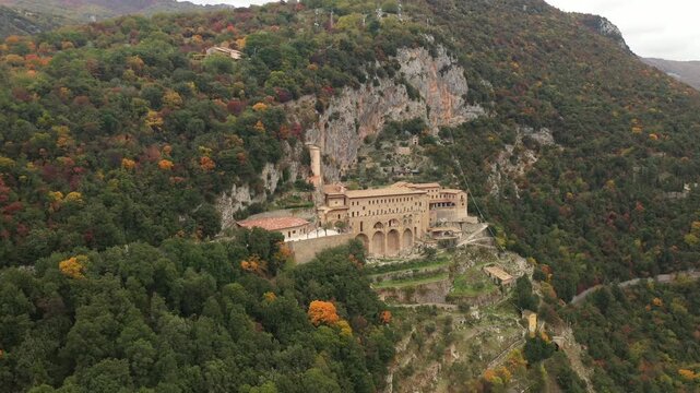 Aerial view of the monastery of St Benedict nestled into the cliffside amidst lush green and autumnal trees, creating a beautiful contrast, Subiaco, Lazio, Italy.