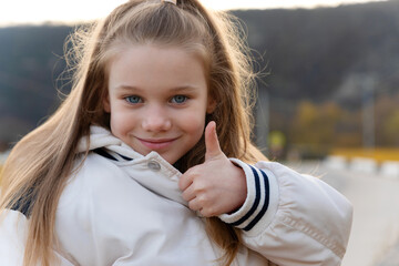 Cheerful young girl in a white jacket showing thumbs up outdoors, expressing positivity and confidence on a sunny day.