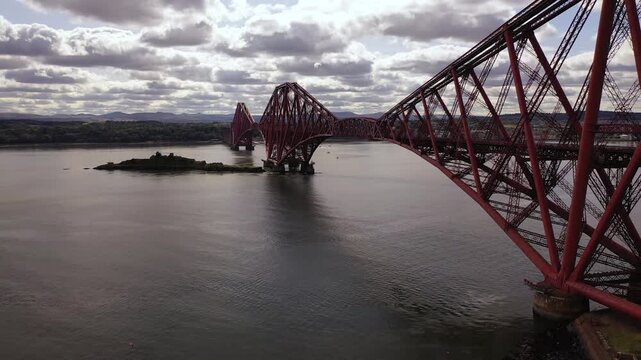 Aerial view of the red Forth Bridge stretching across the still water, with a small island nearby, under a sky of dramatic clouds, Inverkeithing, Scotland, United Kingdom.
