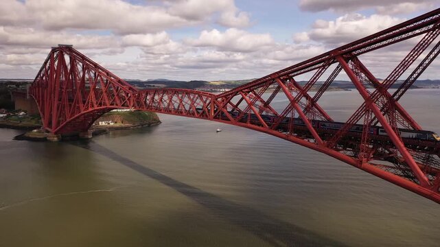Aerial view of the Forth Bridge, its red steel contrasting with the dark water and the green shores, a train crossing, Inverkeithing, Scotland, United Kingdom.