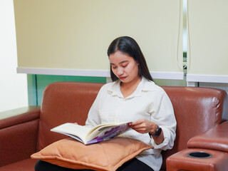 Dark skinned Asian woman sitting on sofa and reading book inside office during her break in work day. Female wearing casual outfit enjoying learning education book relax time. Knowledge. Goal planning