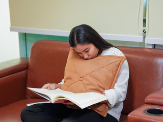 Dark skinned Asian woman sitting on sofa and reading book inside office during her break in work day. Female wearing casual outfit enjoying learning education book relax time. Knowledge. Goal planning