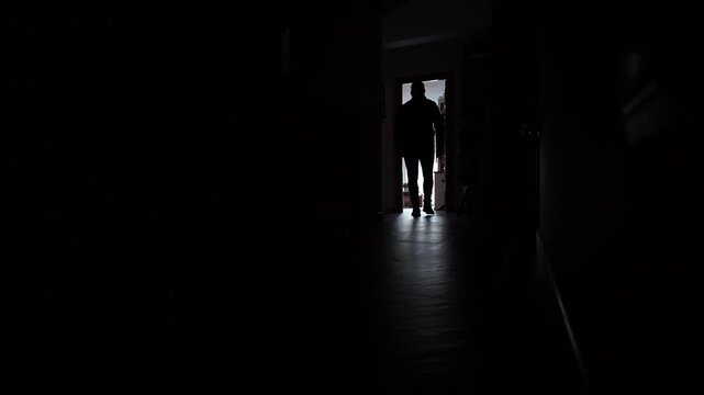 Mysterious male figure walking down a dark corridor towards a bright light coming from a doorway