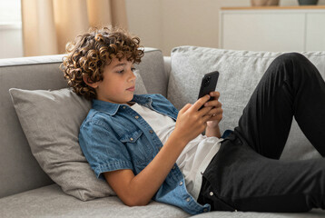 Curly - haired boy using smartphone while relaxing on gray couch at home - childhood technology leisure