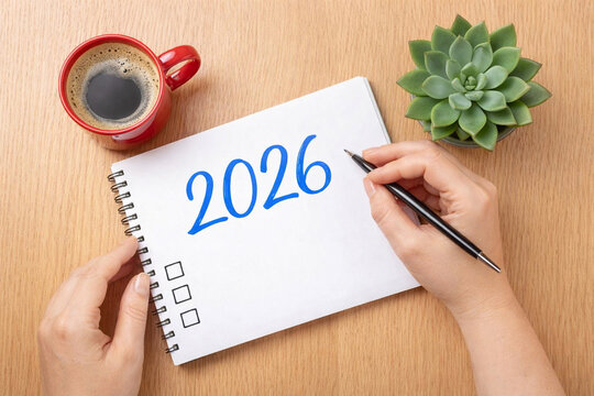 New Year Resolutions 2026 on Wooden Desk, Hands Writing in Notebook, Coffee Cup and Plant, Copy Space