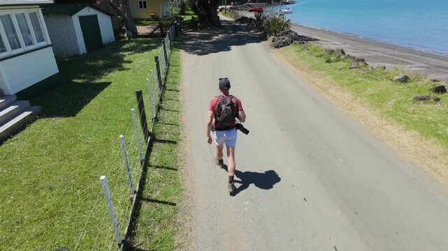 A photographer with a backpack walks along a road next to the beach in Huia, New Zealand. He is likely scouting locations for a photoshoot. The day is sunny and bright.