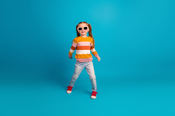 Young girl wearing a colorful striped sweater with sunglass stands playfully against a bright blue background fashion style and joyful childhood