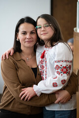 Vertical photo of smiling mother hugging her daughter in vyshyvanka indoors, warm family emotion concept