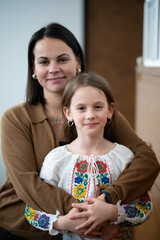 Vertical photo of smiling middle aged mother gently hugging her daughter in vyshyvanka indoors, family warmth emotion concept