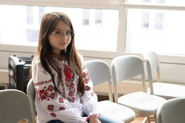 Cute shy schoolgirl in glasses wearing vyshyvanka with long hair sitting on chair in classroom, school education concept