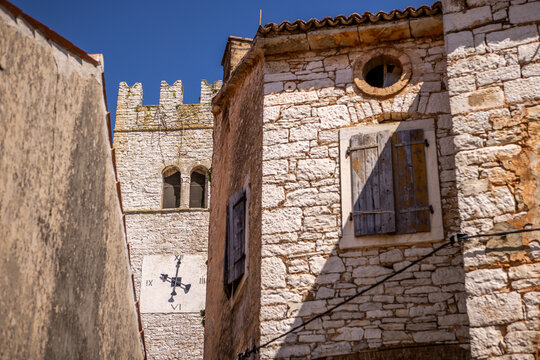 View of sun-drenched stone walls meet weathered wooden shutters and an ancient clock tower under a clear blue sky, Groznjan, Istria County, Croatia.
