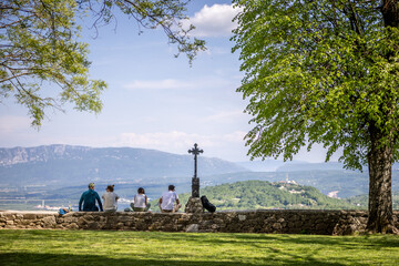 View of a serene vista unfolds from a grassy knoll where figures sit near a stone wall and metal cross, framed by lush trees, Groznjan, Istria County, Croatia.