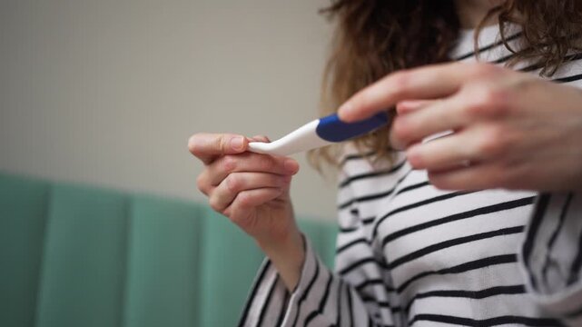 Young woman's hands holding a pregnancy test stick, looking at the positive result with a mix of surprise and emotion. The focus is on the discovery of early motherhood and family planning