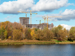 Residential building construction site with high tower cranes under white clouds and blue sky view from autumn riverbank