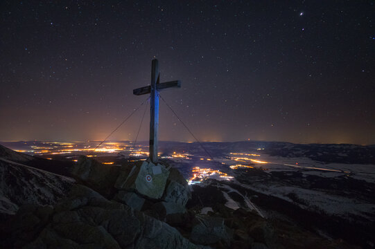 View of a cross atop a rocky peak under a starry sky, with a distant cityscape glowing softly in the night, Vysoke Tatry, Presovsky kraj, Slovakia.