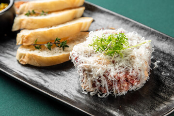 A gourmet beef tartare dish topped with shredded parmesan cheese and fresh greens, served with toasted bread slices on a black plate.