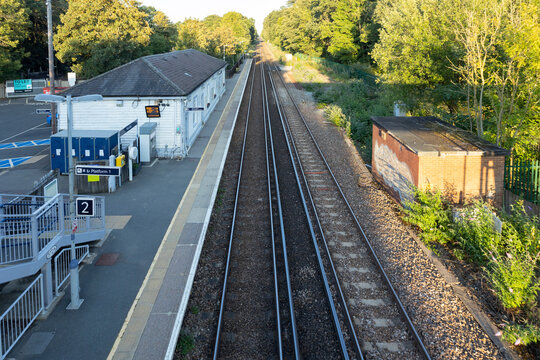Elevated view of a quiet train platform with parallel tracks, a small station building, and lush trees. A calm, everyday transit scene capturing travel,