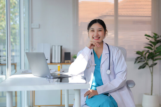 Asian woman doctor wearing a lab coat and stethoscope, smiling while sitting at an office desk with a laptop