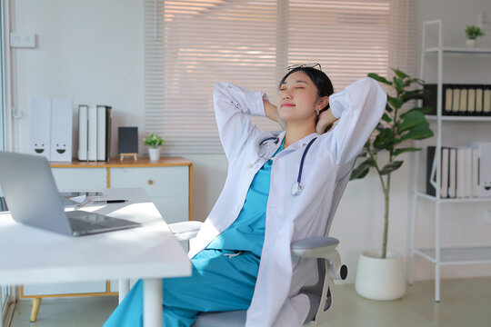 Asian female doctor in scrubs and lab coat, leaning back in an office chair, eyes closed, enjoying a relaxing moment during a break