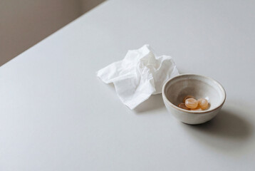 Cough drops in a ceramic bowl beside crumpled tissue on a white table, home cold and flu care concept with minimal still life and copy space