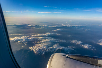 Panoramic aerial view of layered mountains and clouds from high altitude airplane window showing engine cowling and brilliant sunlight reflection.