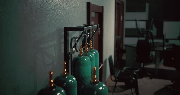 In a dimly lit area of an industrial setting, several green gas cylinders stand in a row, casting shadows on a textured wall. A sparse, utilitarian atmosphere surrounds the space.