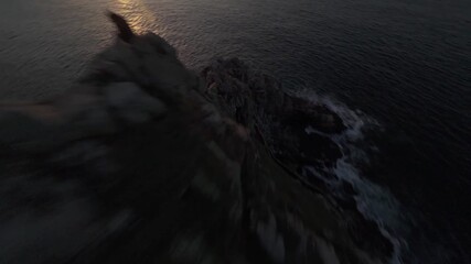 Cinematic drone shot of a rugged rocky headland at sunset, captured in RAW for maximum grading flexibility and rich highlight detail. Warm goldenhour tones, calm coastal mood.
