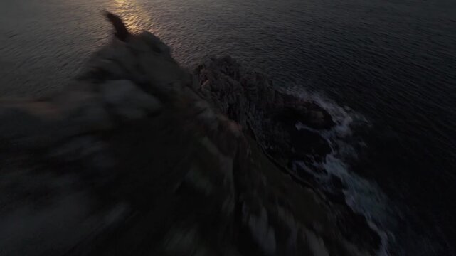 Cinematic drone shot of a rugged rocky headland at sunset, captured in RAW for maximum grading flexibility and rich highlight detail. Warm goldenhour tones, calm coastal mood.