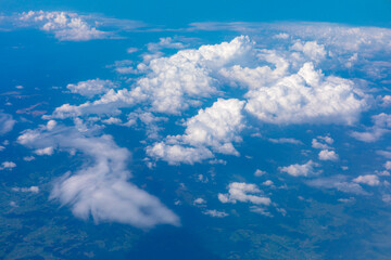 Billowing cumulus clouds drift above a patchwork of green terrain, casting soft shadows over the earth. Aerial view captures the serene contrast between sky texture and land contours