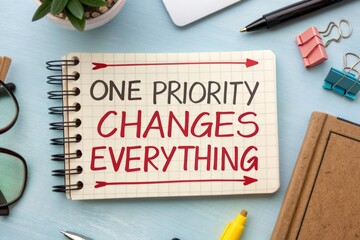 Top down view of a spiral bound notebook with the phrase One Priority Changes Everything written in bold red lettering, surrounded by office supplies