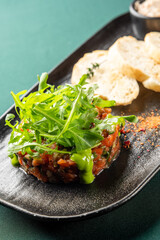 Finely chopped salmon tartare appetizer topped with fresh arugula and green sauce, served with sliced baguette on a black plate