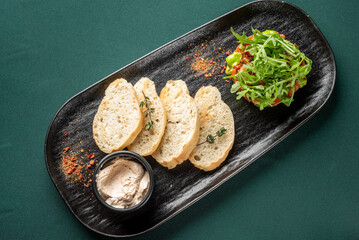 Finely chopped salmon tartare appetizer topped with fresh arugula and green sauce, served with sliced baguette on a black plate