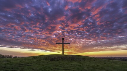 A wooden cross stands atop a grassy hill under a vibrant sunset sky with colorful clouds.