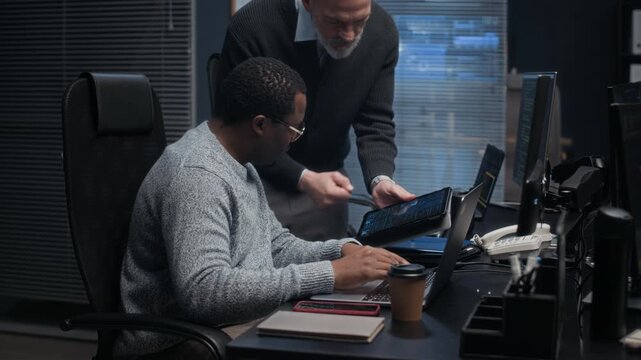 Two ethnically diverse cybersecurity colleagues working late in dark office reviewing data and performing security tasks on digital tablet while monitoring network systems